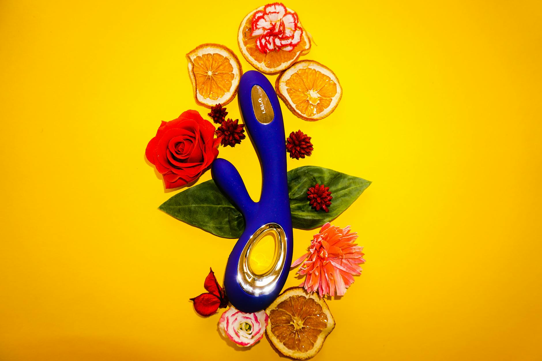 Blue clitoral vibrator surrounded by flowers and citrus on bright yellow background